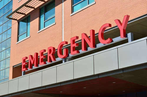Red lettering signage above a grey hospital building canopy, spelling the word EMERGENCY