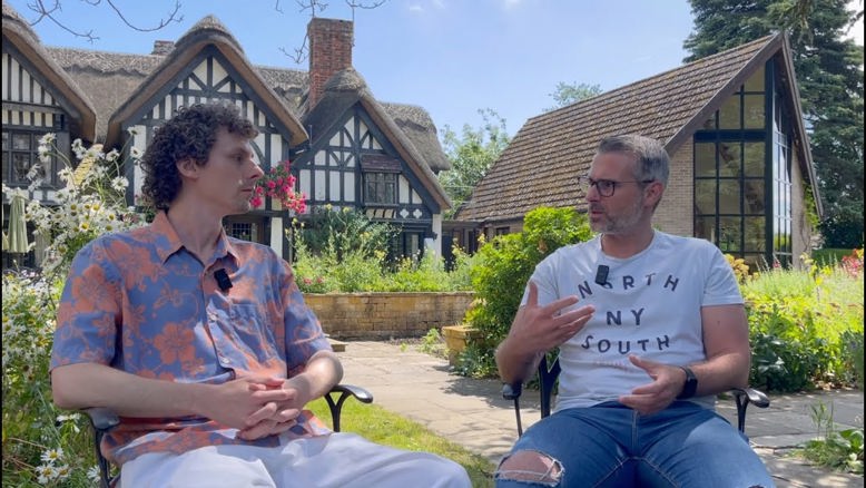 Two people sitting down looking at each other and conversing, in front of greenery and a Tudor style building in black and white